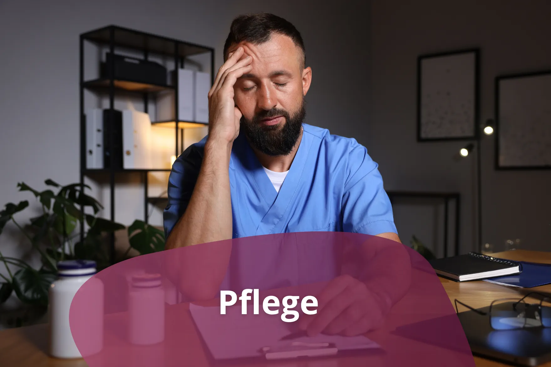 Erschöpfte männliche Pflegekraft in blauem Kasack sitzt mit geschlossenen Augen am Schreibtisch und hält sich die Stirn.