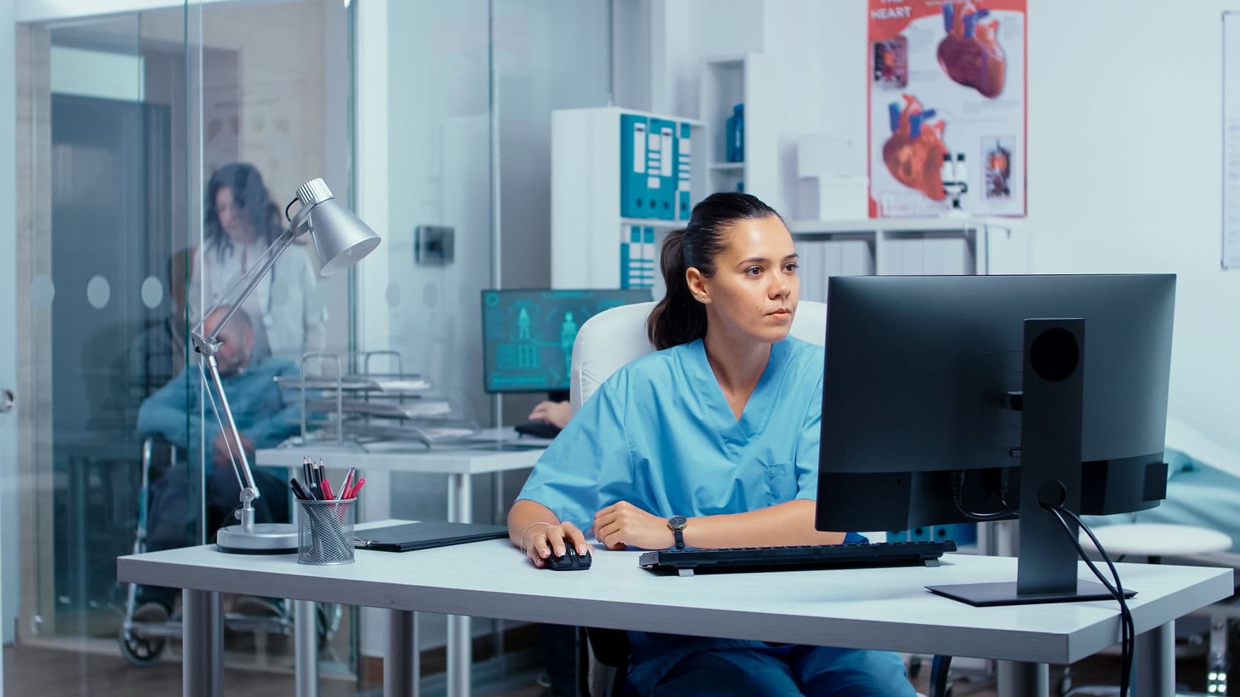 Krankenschwester mit dunklen Haaren und Zopf sitzt im Krankenhaus in einem Büro und arbeitet am PC.