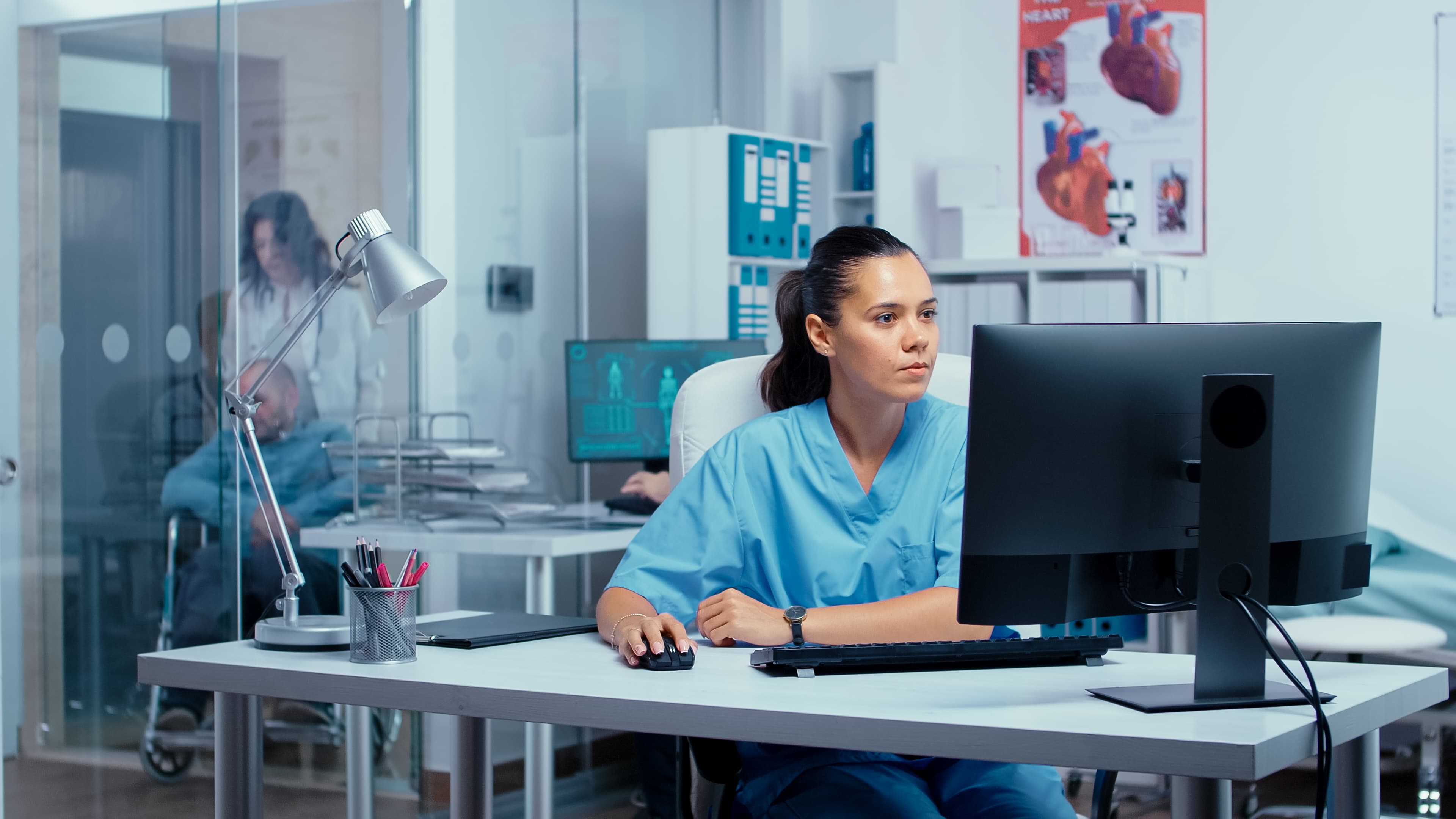 Krankenschwester mit dunklen Haaren und Zopf sitzt im Krankenhaus in einem Büro und arbeitet am PC.