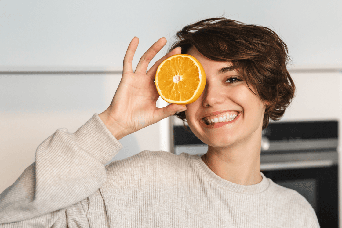 Lächelnde junge Frau verdeckt ein Auge mit einer aufgeschnittenen Orange.