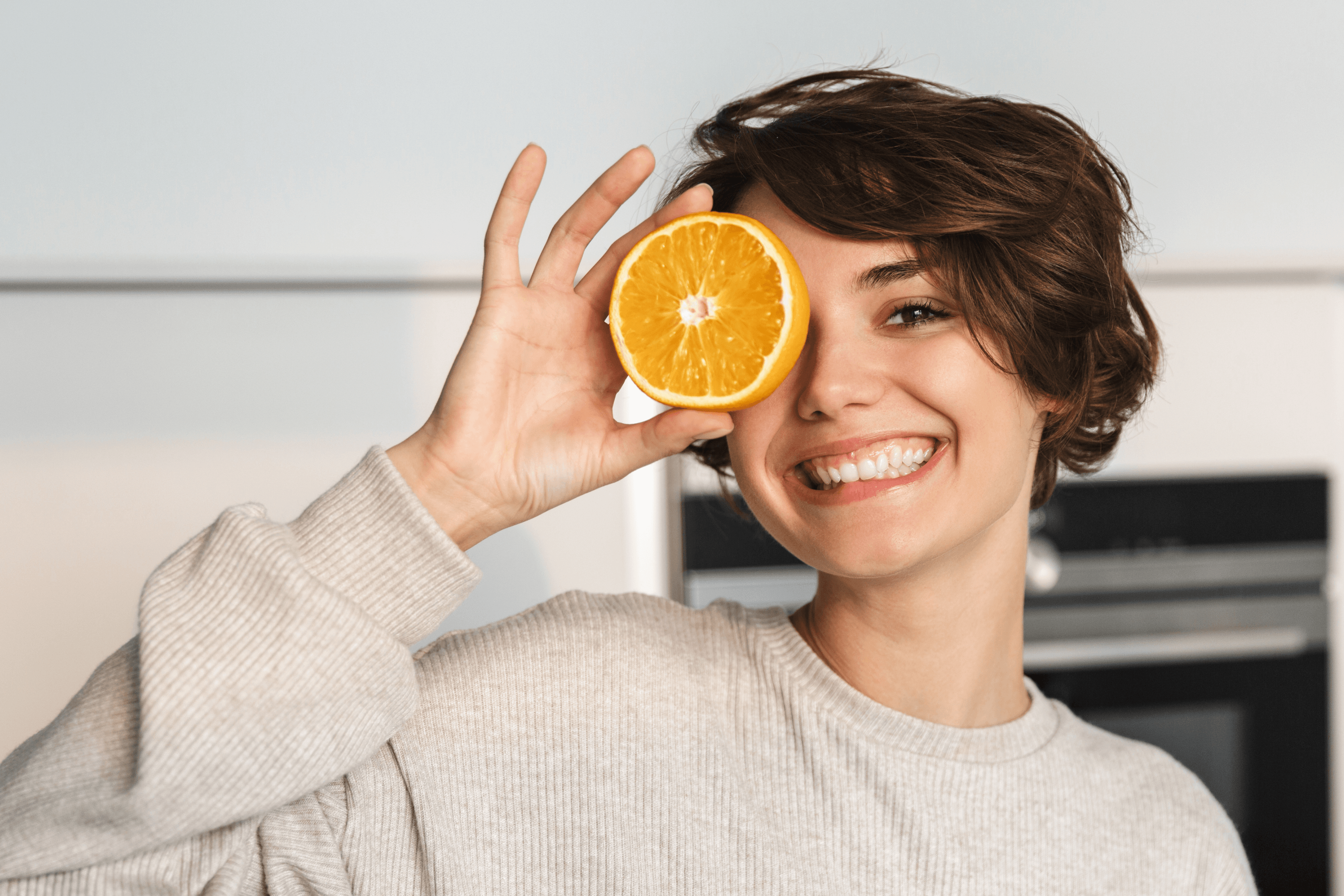 Lächelnde junge Frau verdeckt ein Auge mit einer aufgeschnittenen Orange.