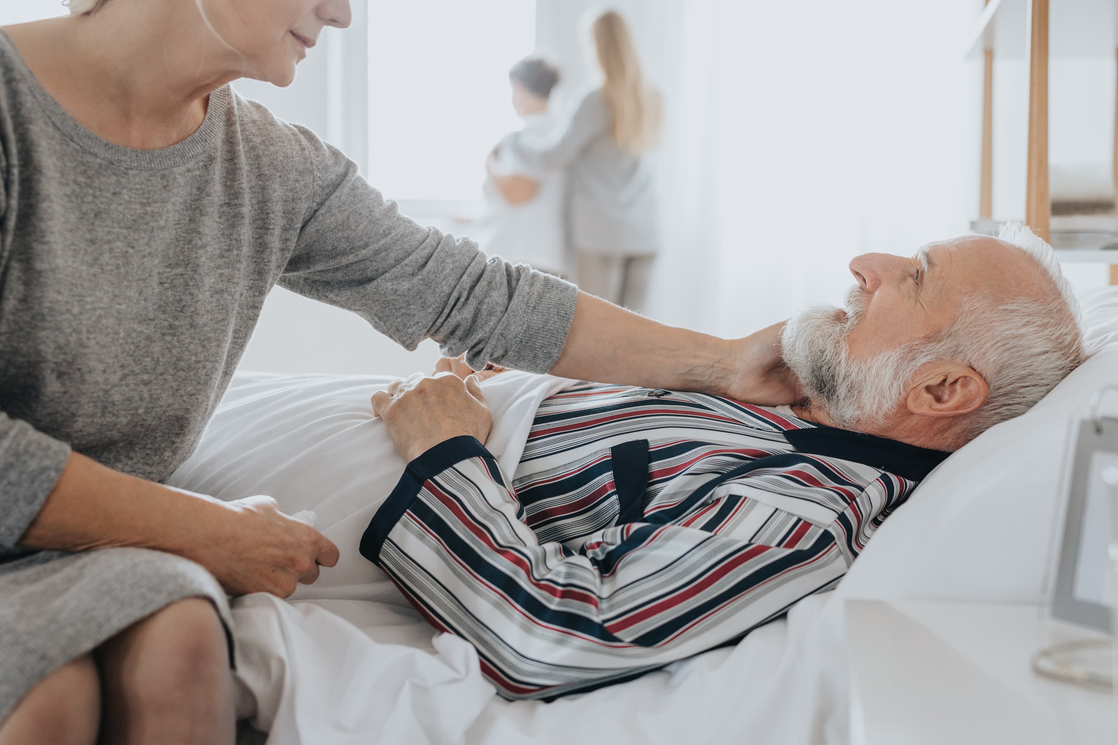 Patient im Bett, seine Frau sitzt am Bettrand und blickt zu ihm