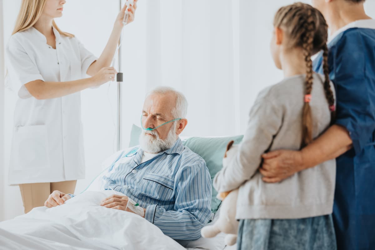 Älterer Mann liegt im Krankenhausbett mit Sauerstoffgerät, wird von Pflegekraft betreut, Familie steht am Bett.