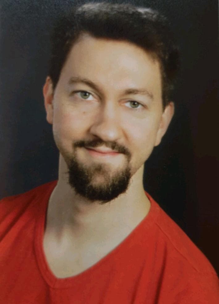 Smiling man with short hair and a beard, wearing a red shirt, against a dark background.