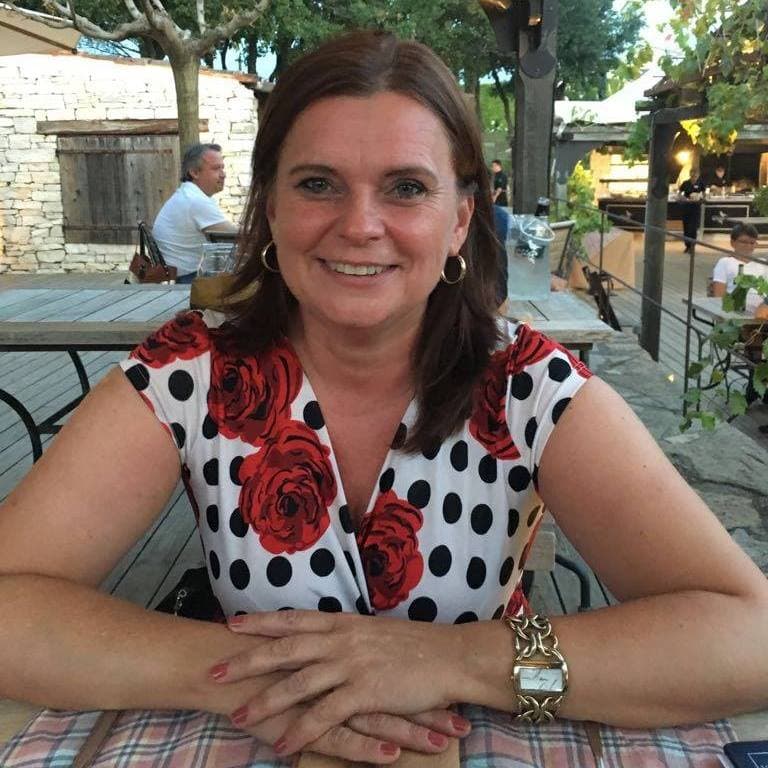 Lächelnde Frau mit braunem Haar sitzt an einem hölzernen Tisch im Freien, trägt ein weiß-schwarz gepunktetes Kleid mit roten Rosen.