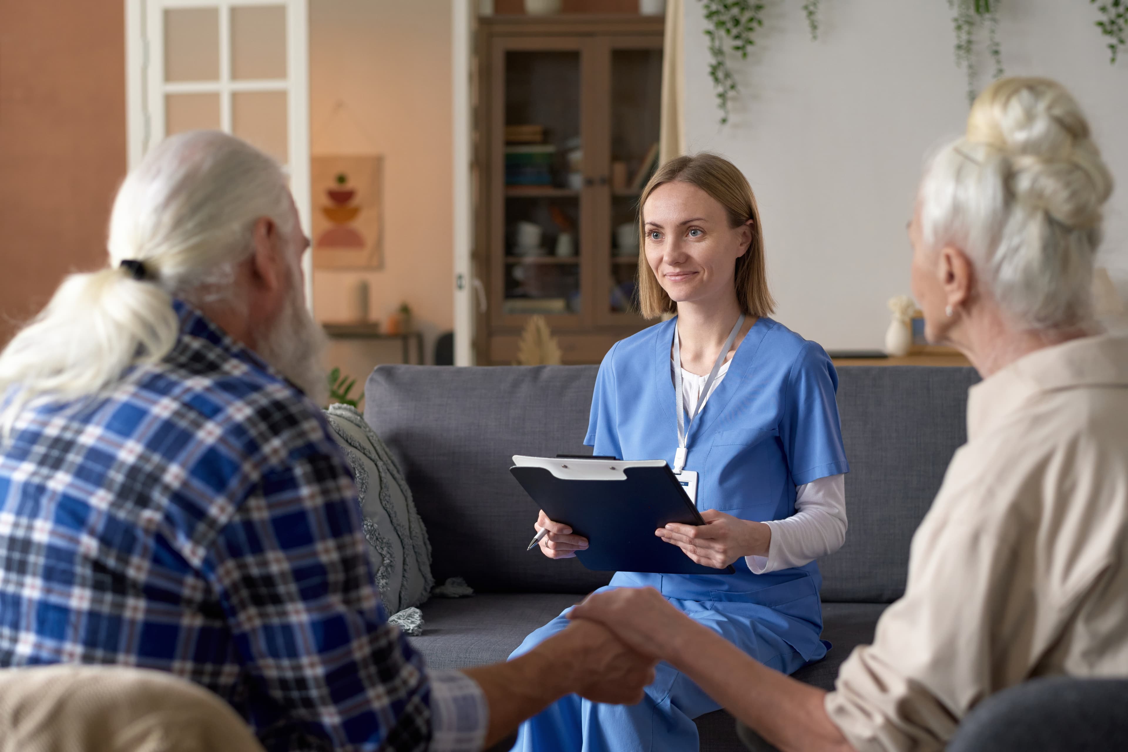 Eine Krankenschwester in blauer OP-Kleidung hält ein Klemmbrett in der Hand und sitzt mit einem älteren Ehepaar auf einem Sofa, die sich in einem gemütlichen Wohnzimmer unterhalten.
