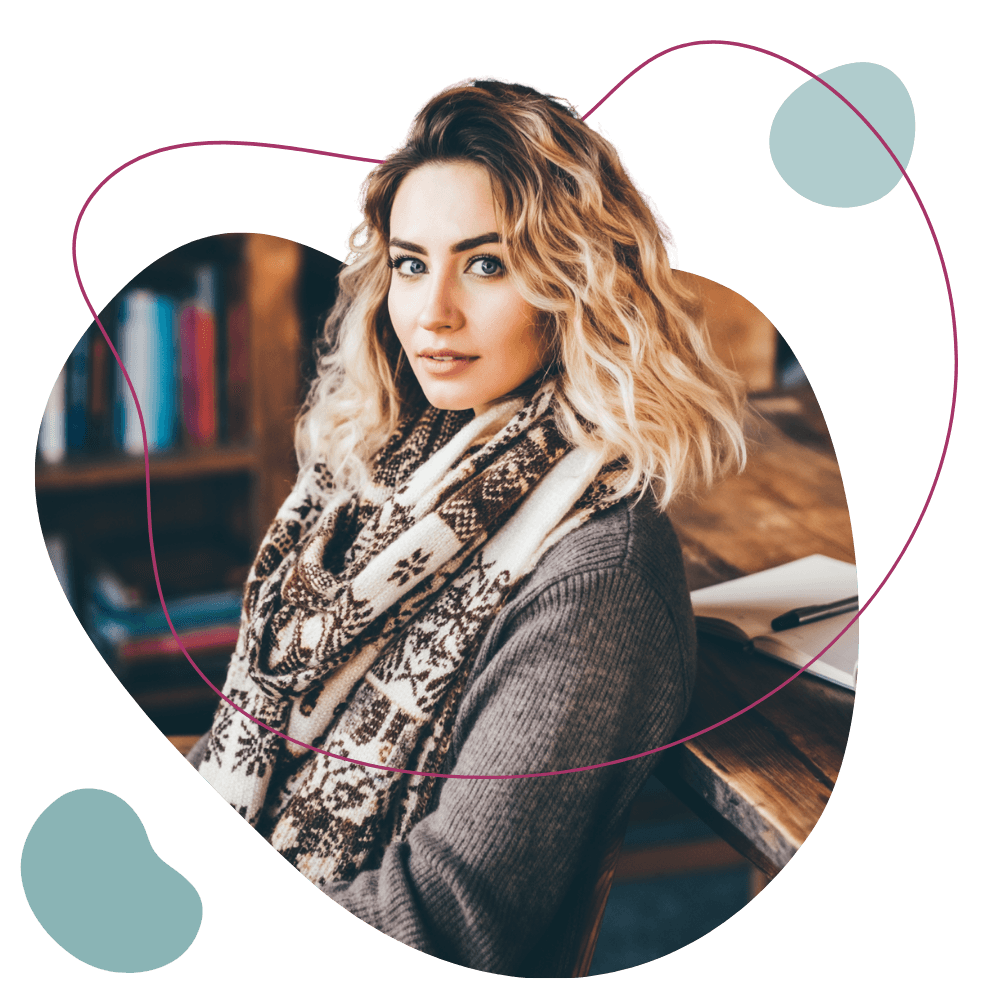 Eine Frau mit einem winterlichen Schal sitzt in ihrem Arbeitszimmer und blickt in die Kamera, Bücher im Hintergrund