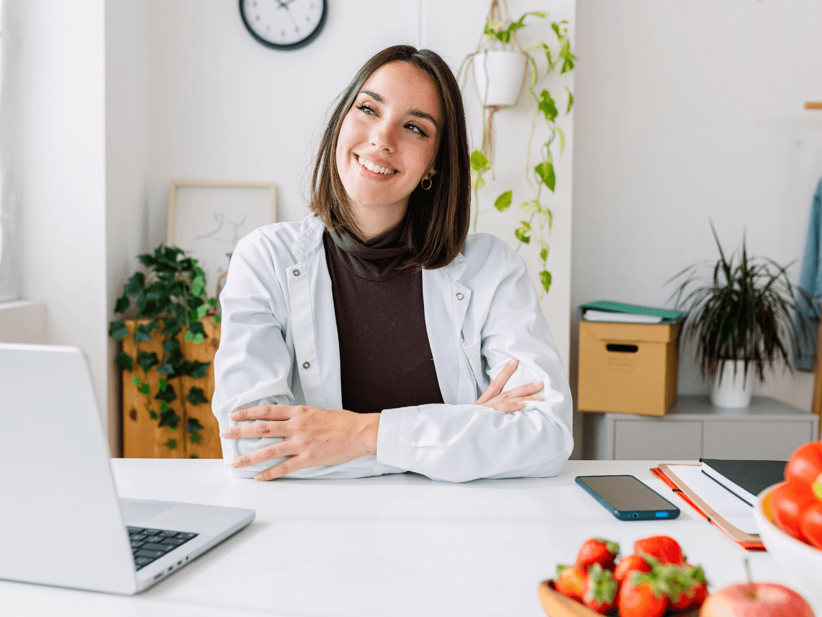 Eine lächelnde Frau im weißen Kittel sitzt an einem Schreibtisch mit Laptop, Telefon und Obst. Im Hintergrund sind Pflanzen und eine Uhr zu sehen.