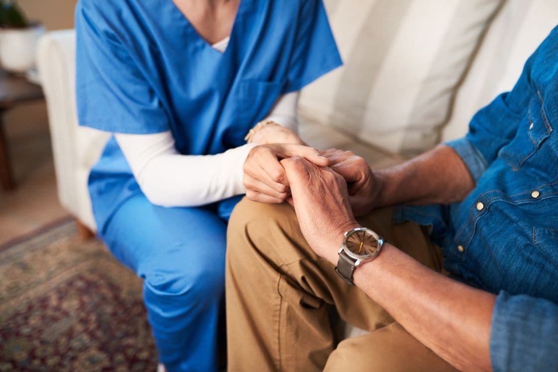 Fachkraft für Palliative Care sitzt mit einem Familienangehörigen auf der Couch und hält seine Hand.
