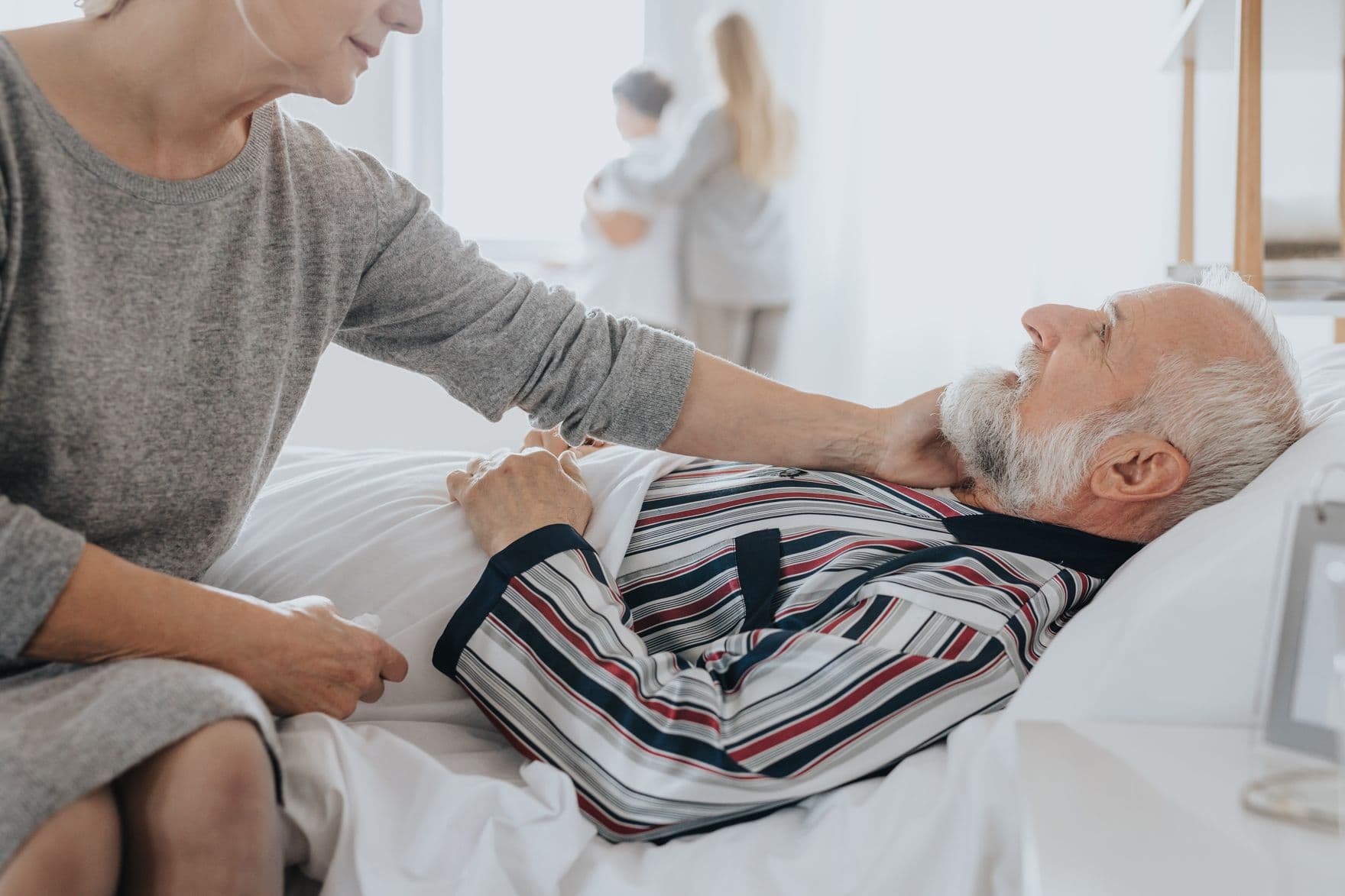 Patient im Bett, seine Frau sitzt am Bettrand und blickt zu ihm